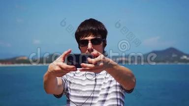 戴太阳镜的年轻人带着他的智能手机在海滩上自拍，用耳机听音乐。 苏梅岛
